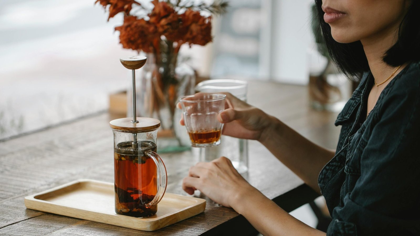 Person enjoying tea at a rustic cafe, with a French press and glass cup on a wooden table.