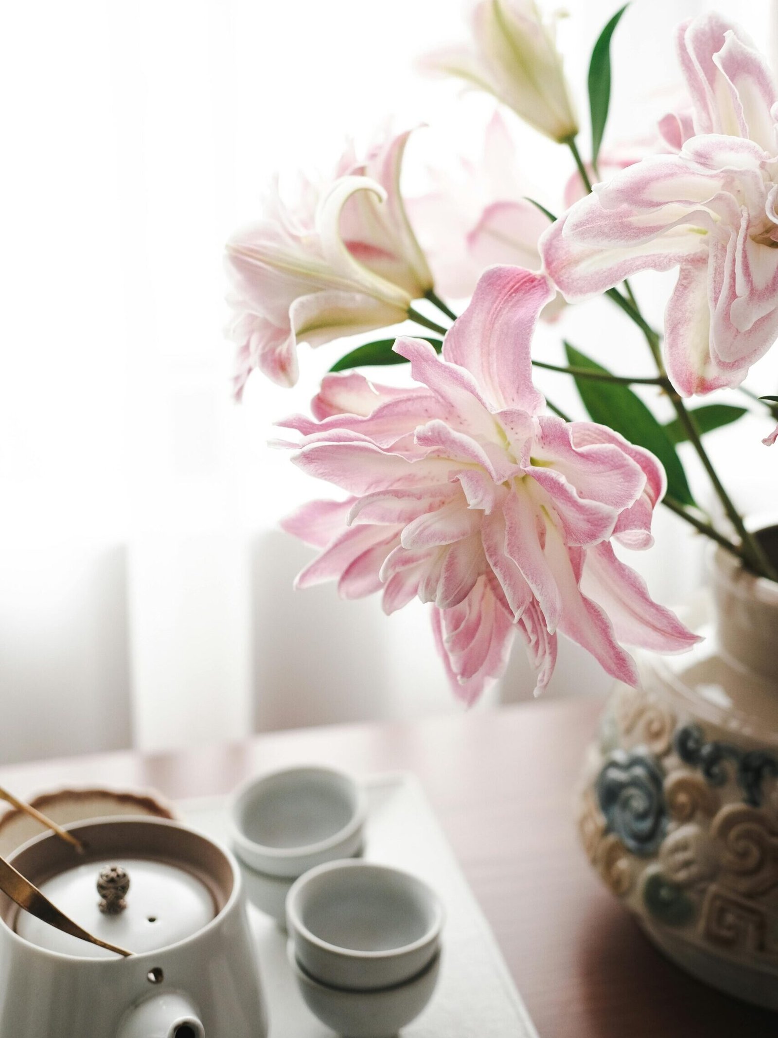 Pink lilies in a decorative vase beside a tea set on a wooden table, bathed in soft natural light.