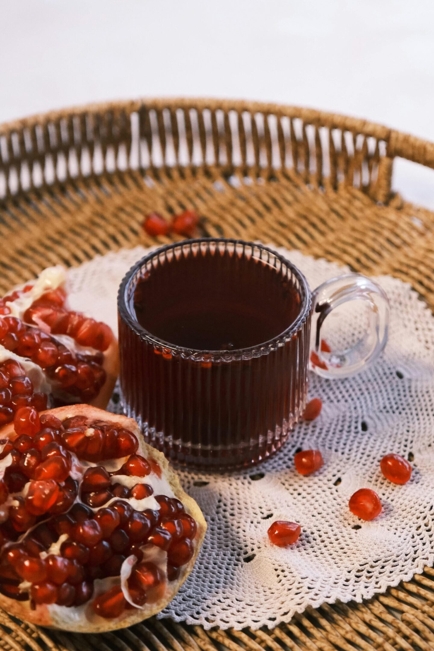 Glass of pomegranate juice with fresh pomegranate seeds on a woven tray and lace doily for a cozy setting.