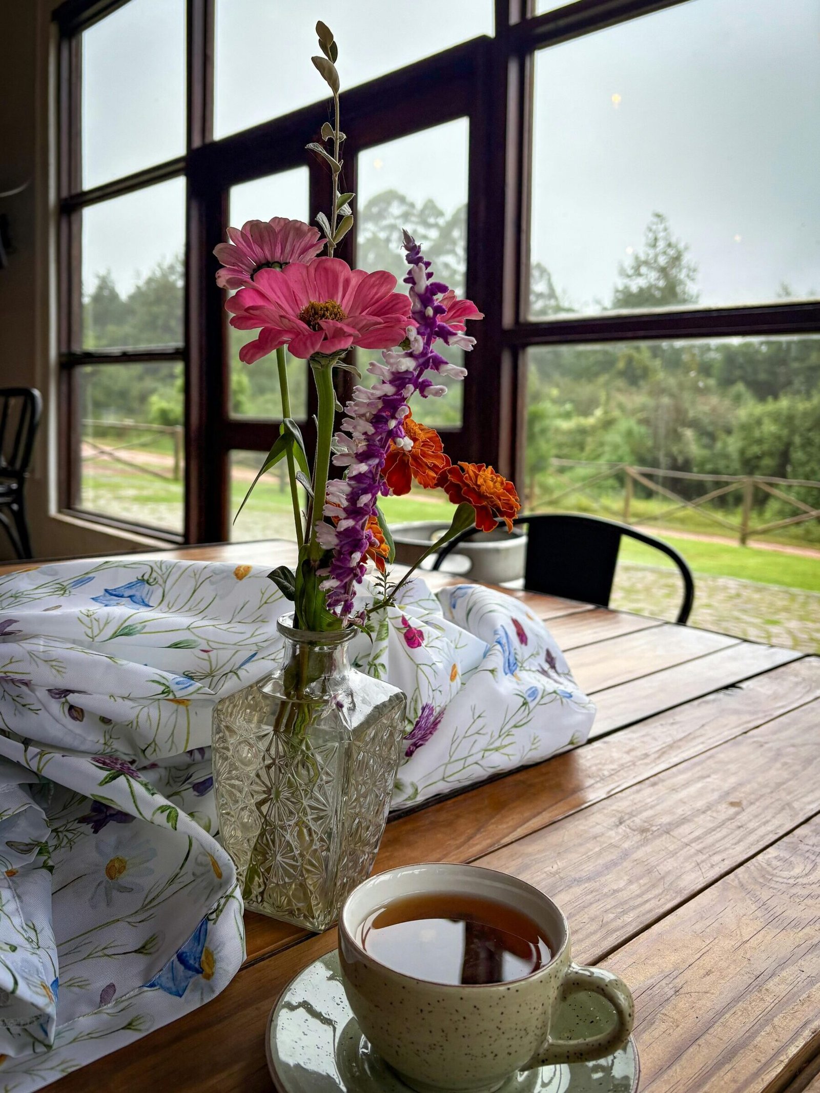 Cozy café scene with flowers in a vase and a steaming cup of tea on a wooden table by a large window.