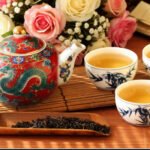 Ornate Chinese teapot with dragon and floral design, accompanied by three cups of tea and fresh roses.