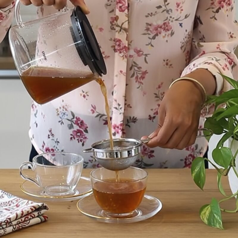 Person pouring tea through strainer into glass cup on wooden table with floral decor.