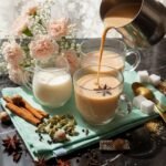 Pouring chai tea into glass cups with spices and milk on a table, next to sugar cubes and pink flowers.