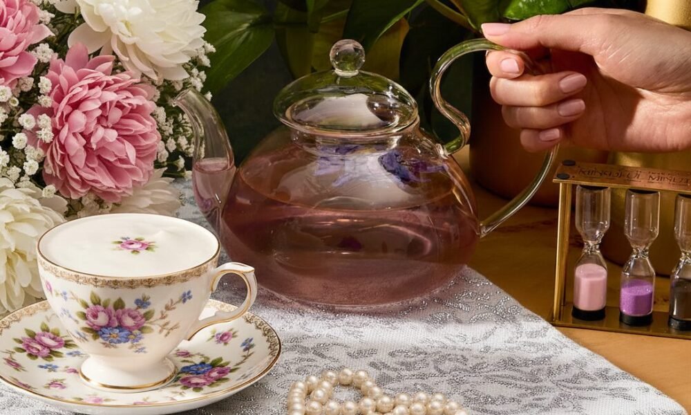 Elegant tea setup with floral cup, glass teapot, blooming bouquet, pearls, and sand timer on lace tablecloth.
