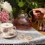 Elegant tea setup with floral cup, glass teapot, blooming bouquet, pearls, and sand timer on lace tablecloth.
