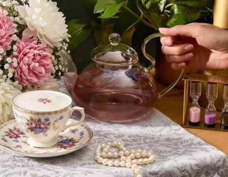 Elegant tea setup with floral cup, glass teapot, blooming bouquet, pearls, and sand timer on lace tablecloth.