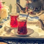 Elegant tea setting with two glasses of red tea and a silver teapot, adorned with fresh flowers on a tray.