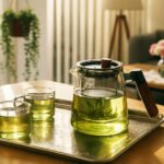 Glass teapot and cups of green tea on a tray, with sunlight streaming into a cozy living room.