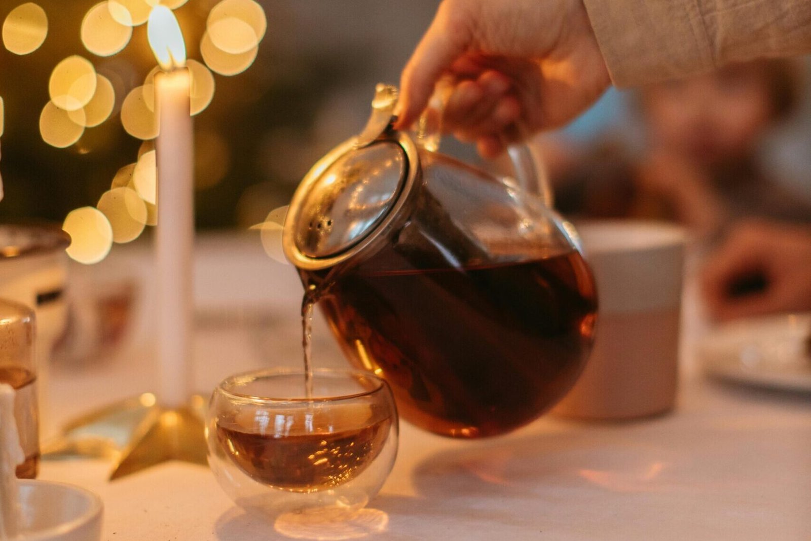 Pouring tea from a glass teapot into a double-walled cup with a cozy, candlelit background.