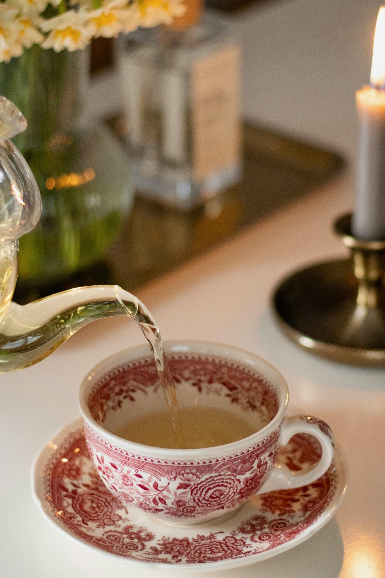 Pouring tea into a vintage teacup, surrounded by flowers and a lit candle for a cozy ambiance.