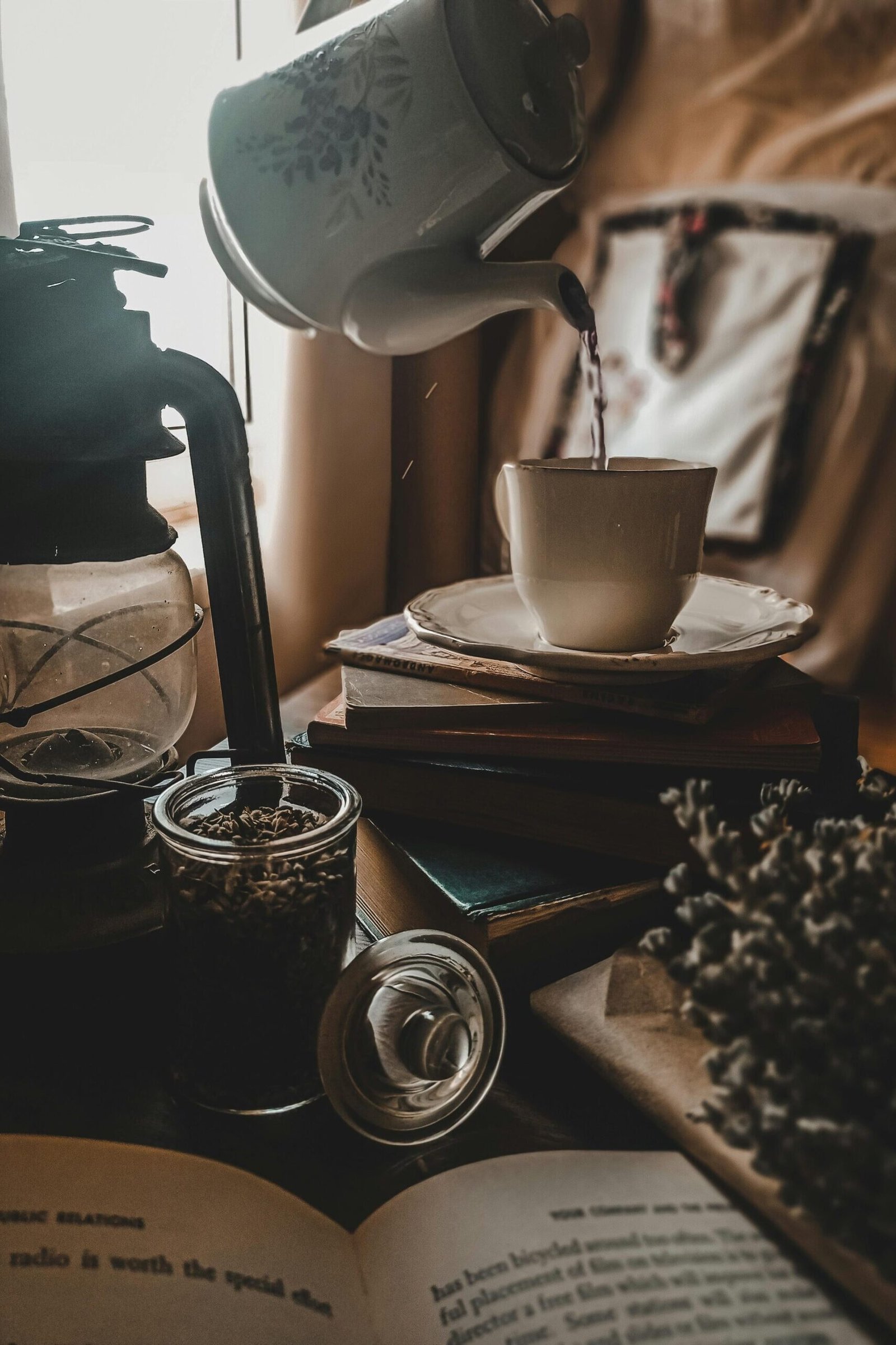 Cozy scene with tea being poured into a cup, surrounded by books, a lamp, and dried lavender.