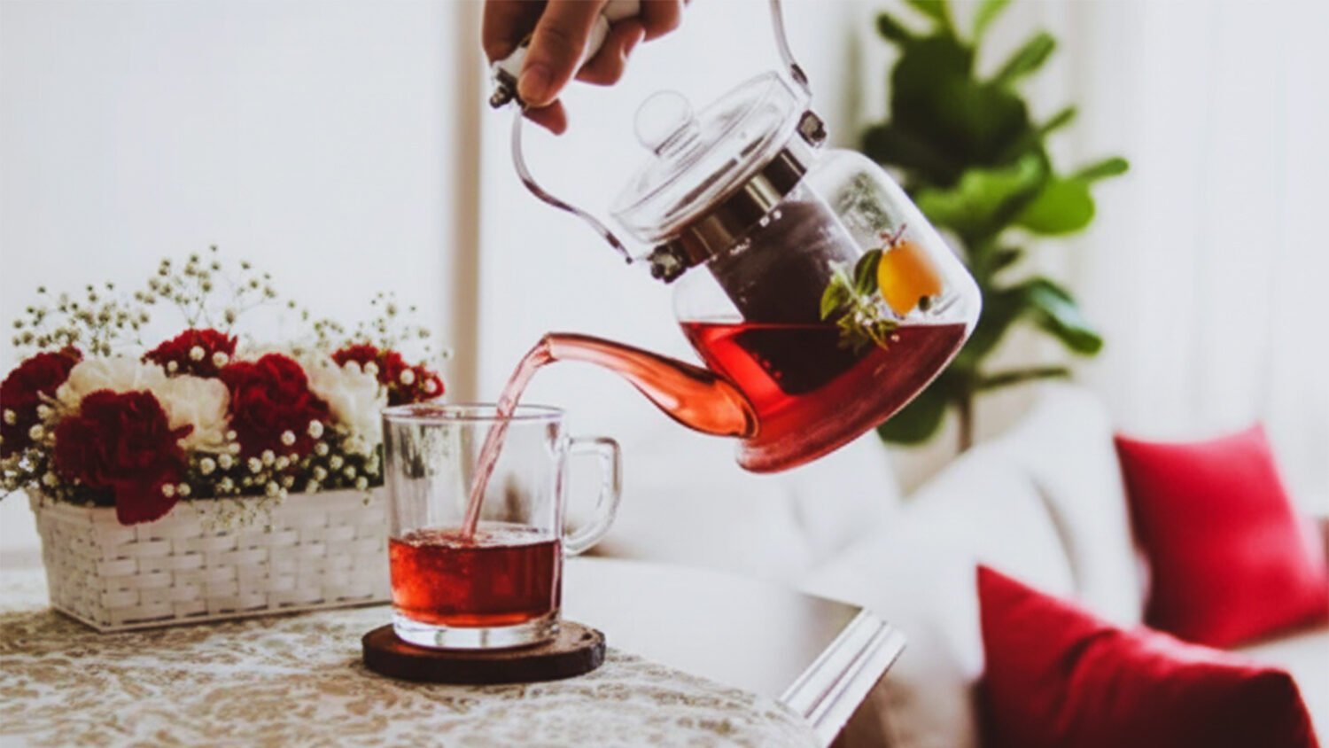 Pouring red tea into a glass cup on a cozy table with floral decor and soft cushions in the background.