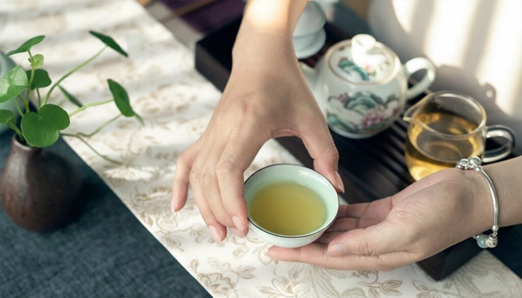Hands holding a green tea cup beside a teapot and plant on a floral tablecloth.