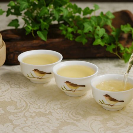 Pouring white tea into bird-themed cups with teapot, accompanied by fresh leaves and tea bowl on patterned cloth.