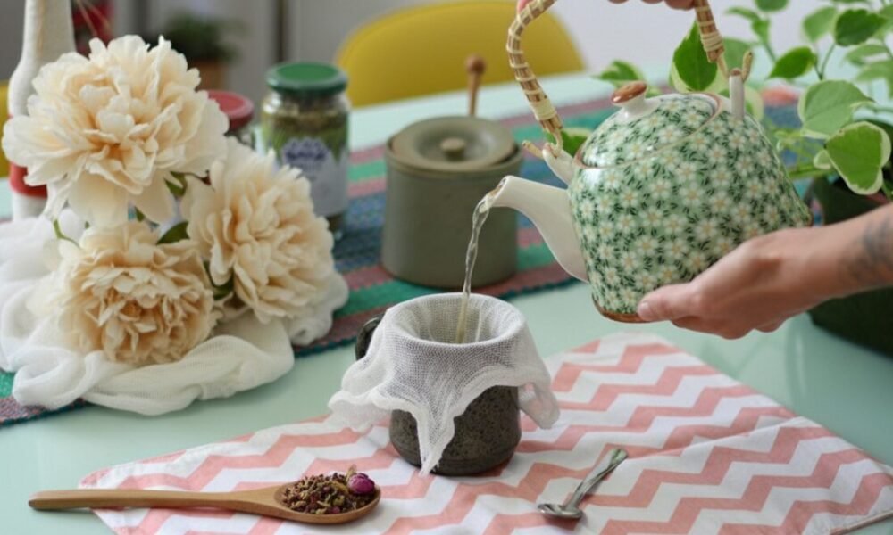 How to Brew Tea Without a Strainer: Easy Loose Leaf Infusions Pouring tea from a floral teapot into a cup, surrounded by flowers and herbs on a patterned cloth.