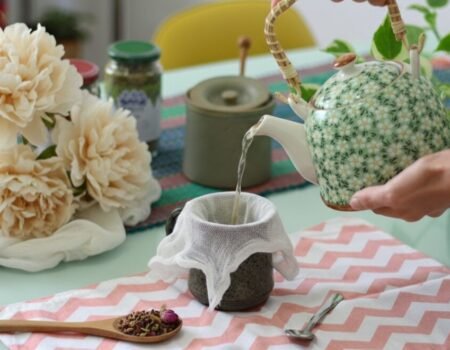 Pouring tea from a floral teapot into a cup, surrounded by flowers and herbs on a patterned cloth.