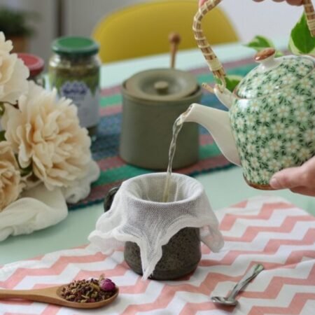 How to Brew Tea Without a Strainer: Easy Loose Leaf Infusions Pouring tea from a floral teapot into a cup, surrounded by flowers and herbs on a patterned cloth.