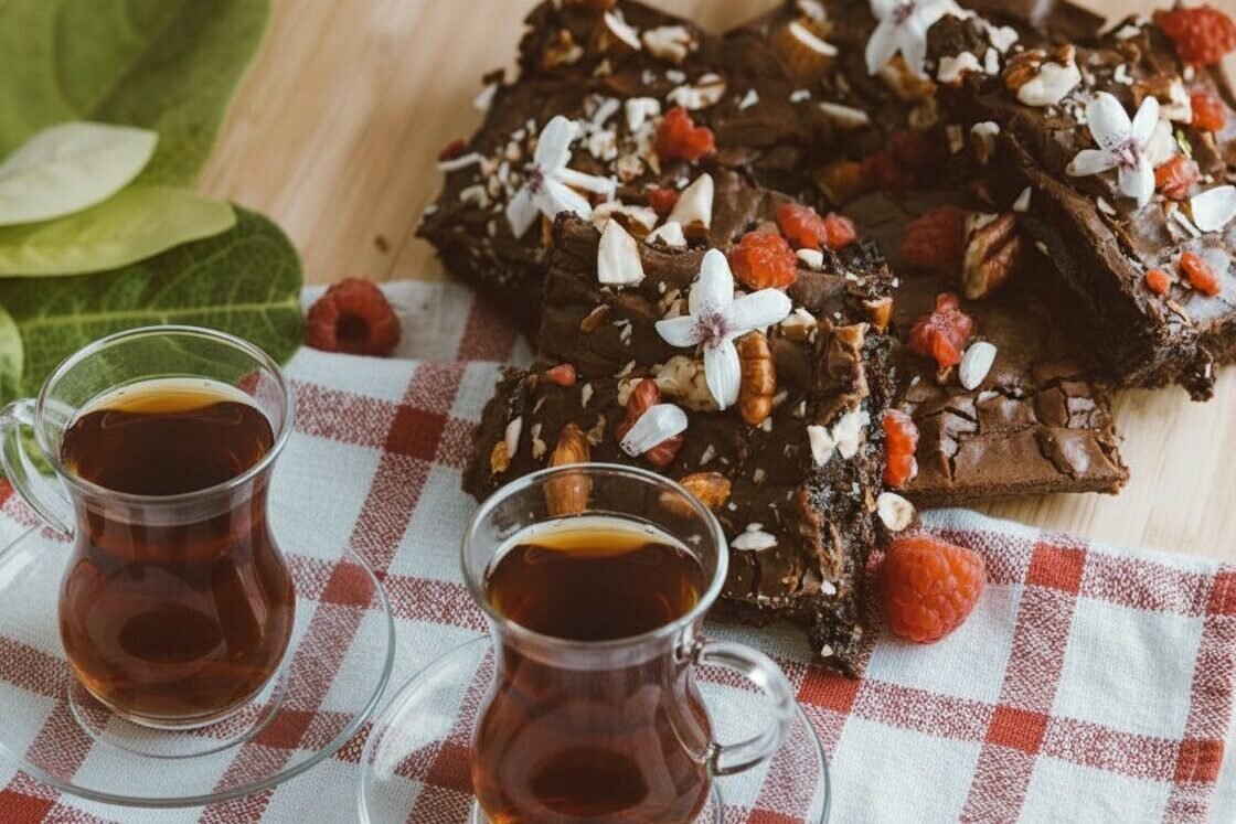 Turkish tea with chocolate brownies topped with nuts and flowers on checkered napkin, surrounded by fresh raspberries.