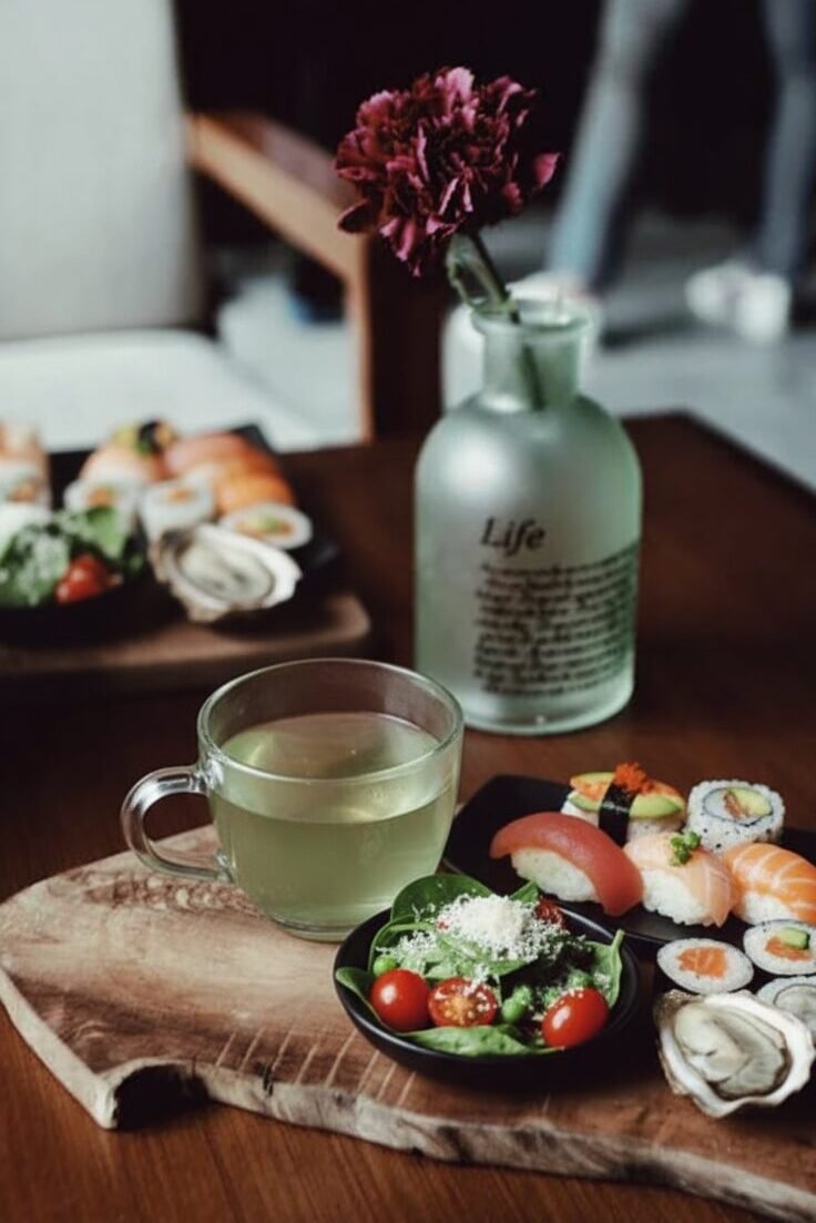 Plate of sushi with a fresh green salad and tea on a wooden board, beside a vase with a red flower.
