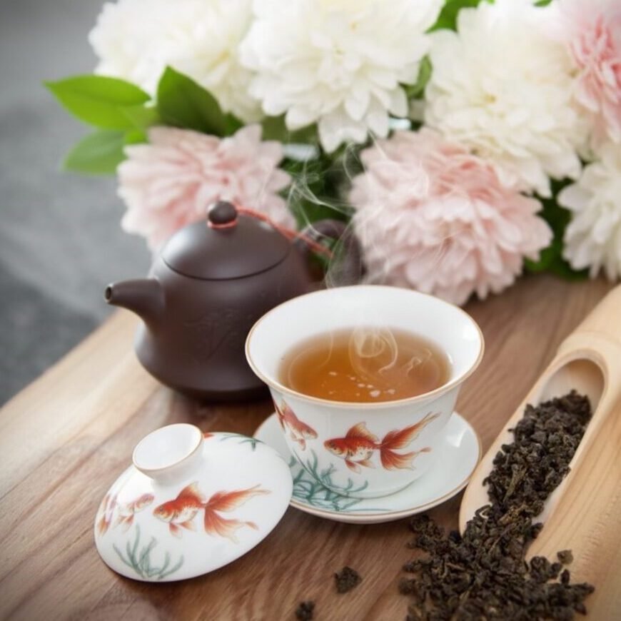 Steaming cup of tea and teapot with floral decor, surrounded by white and pink flowers on a wooden table.