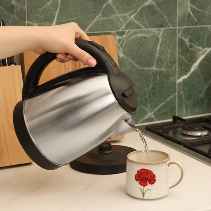 Hand pouring hot water from electric kettle into decorated mug on kitchen countertop with green tile backsplash.