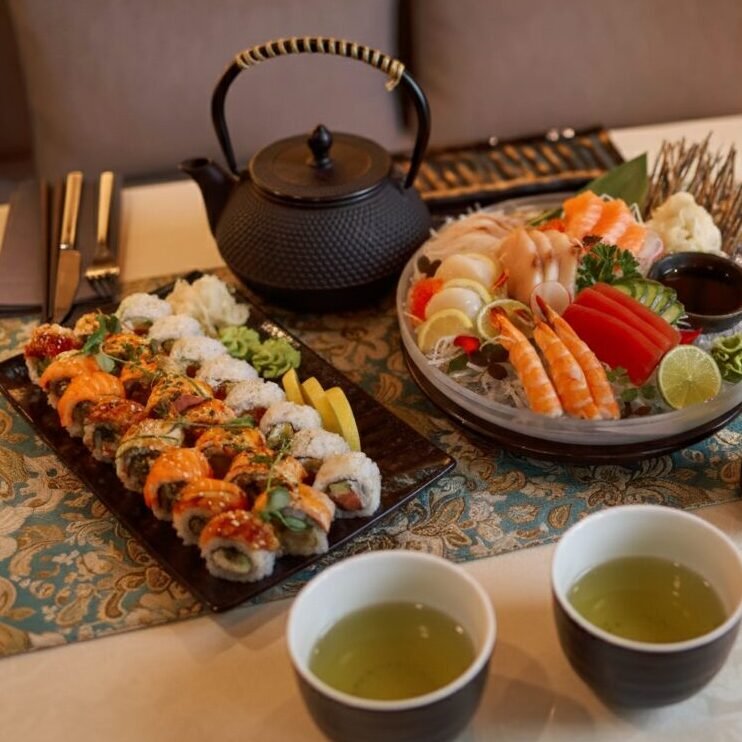 Assorted sushi and sashimi platter with tea set on a patterned tablecloth in a cozy dining setting.
