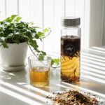 Glass of herbal tea and tea infuser bottle on white table, next to potted plant. Sunlight casts striped shadows.