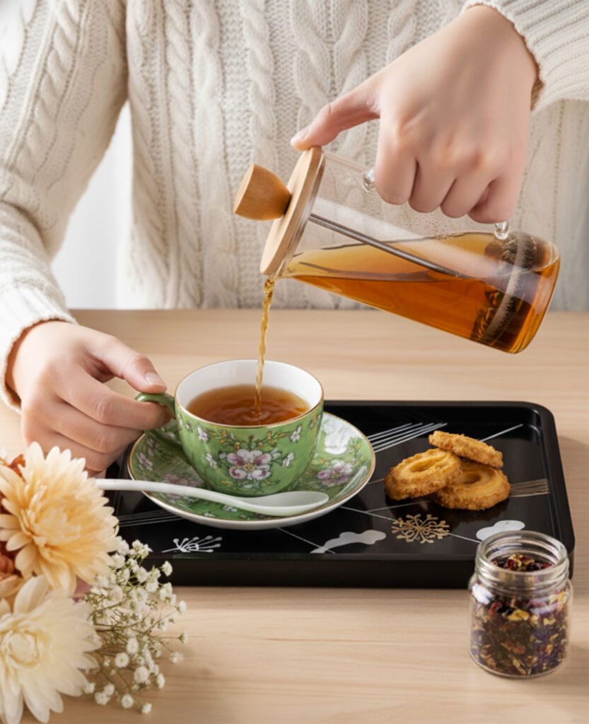 Person pouring tea into floral teacup on tray with cookies, next to flowers and tea jar. Cozy atmosphere.