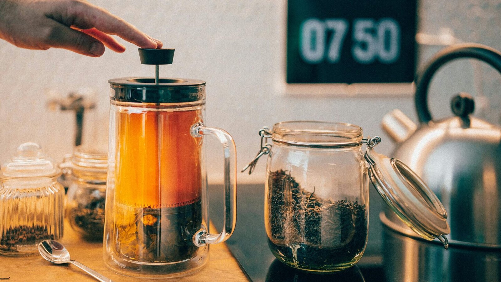 Hand pressing French press with tea near glass jars and kettle on kitchen counter.