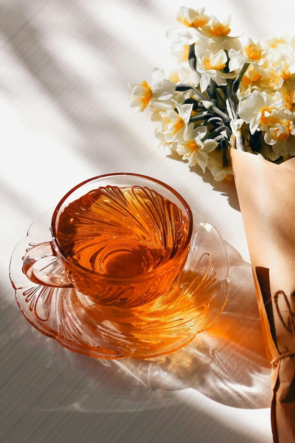 Amber glass teacup on saucer beside a bouquet of daffodils wrapped in brown paper on a sunlit table.
