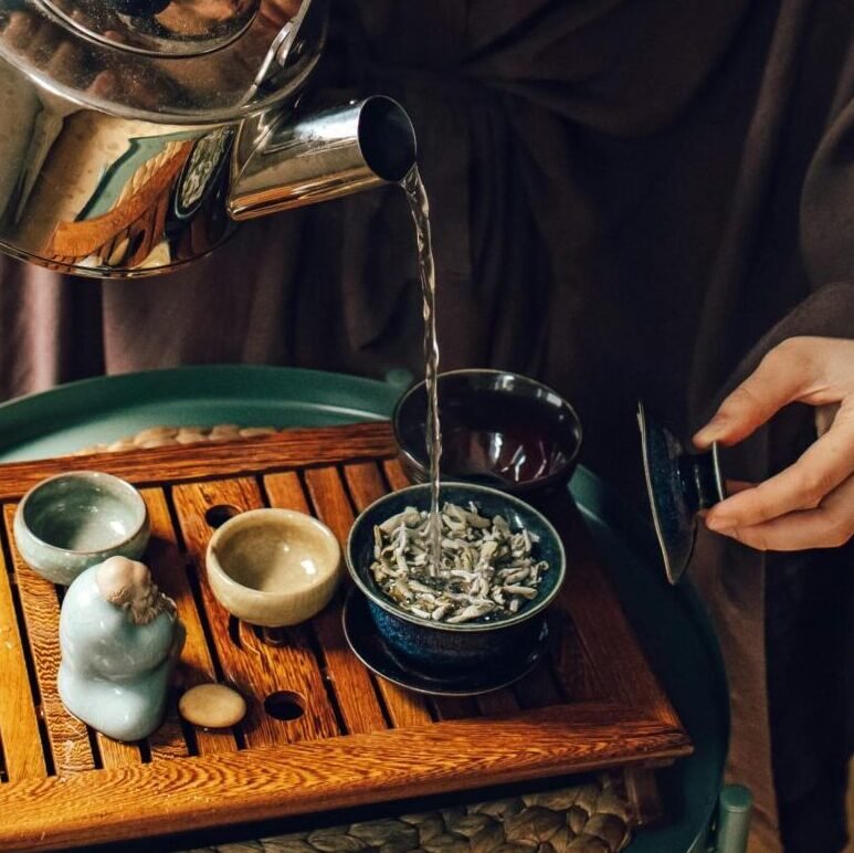 Woman pouring tea from a kettle at home, surrounded by books and plants, creating a cozy atmosphere.