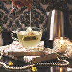 Hand dipping tea bag into cup, with candles, books, and a succulent on a decorative table setup.