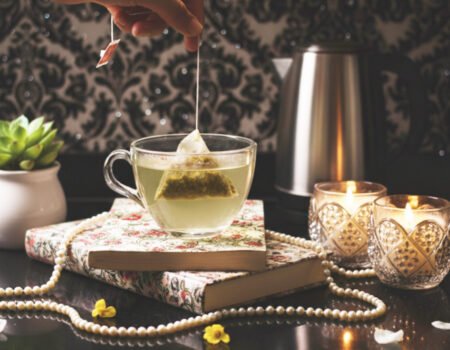 Hand dipping tea bag into cup, with candles, books, and a succulent on a decorative table setup.