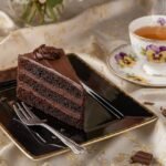 Chocolate cake slice on a black plate with floral teacup on elegant patterned tablecloth.