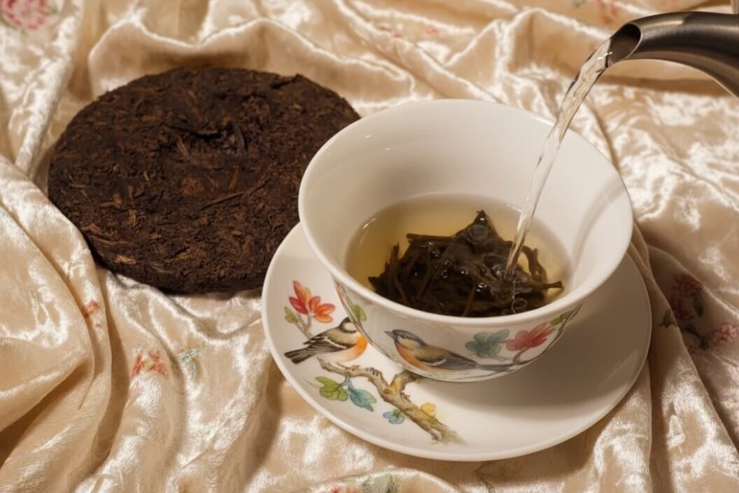 Pouring hot water into a floral teacup with loose leaf tea and a compressed tea cake on silk fabric.