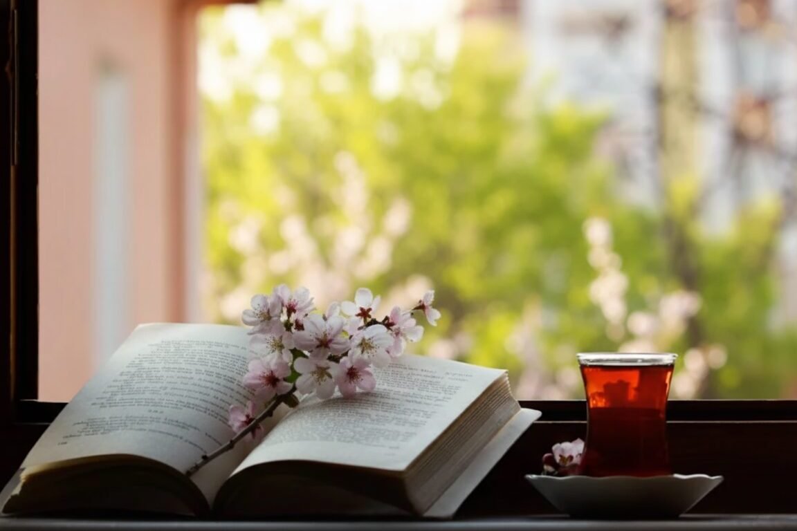 Open book with cherry blossoms and a glass of tea on a windowsill, overlooking a green garden.