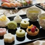 Japanese sweets and sushi on elegant black trays with floral teacups, accompanied by cherry blossoms.