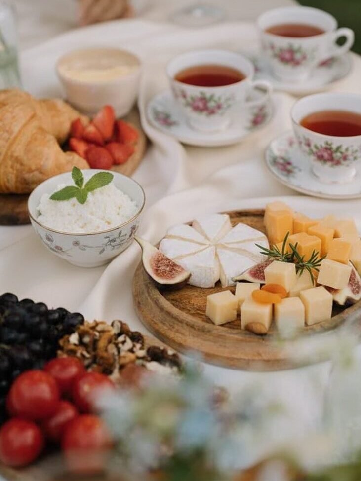 Elegant brunch setting with cheese platter, fruit, croissants, and floral teacups on a white tablecloth.