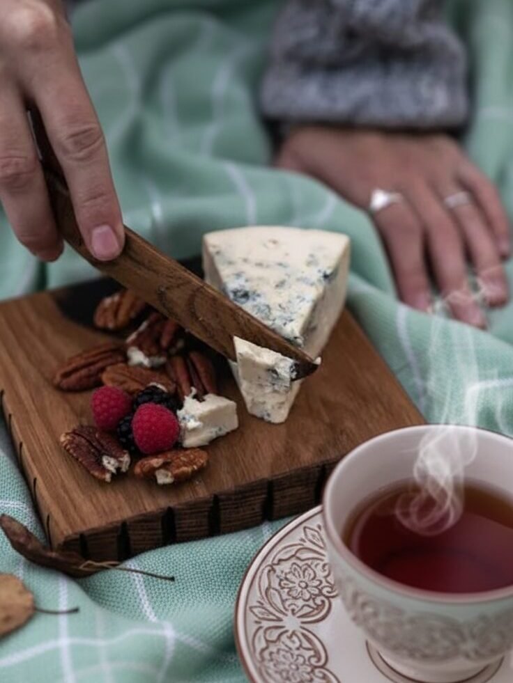 Hand slicing blue cheese with nuts and berries on wooden board, next to steaming cup of tea on a checkered blanket.