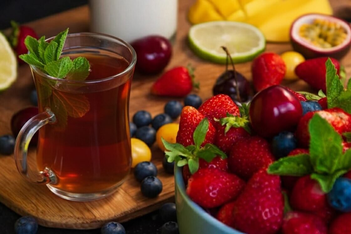 Fresh fruit and herbal tea on a wooden board, surrounded by strawberries, blueberries, cherries, and mint leaves.