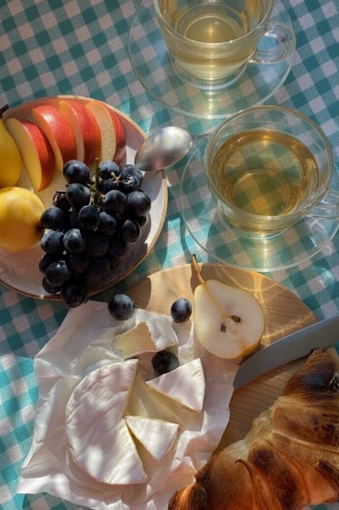 Cheese, pear, croissant, grapes, apple slices, tea on picnic table with blue checkered cloth basking in sunlight.