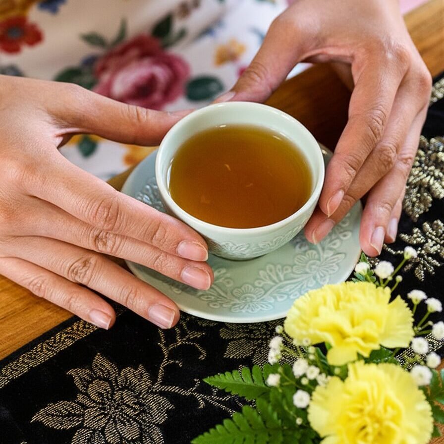 Hands holding a teacup on a patterned tablecloth with yellow flowers, capturing a serene tea moment.
