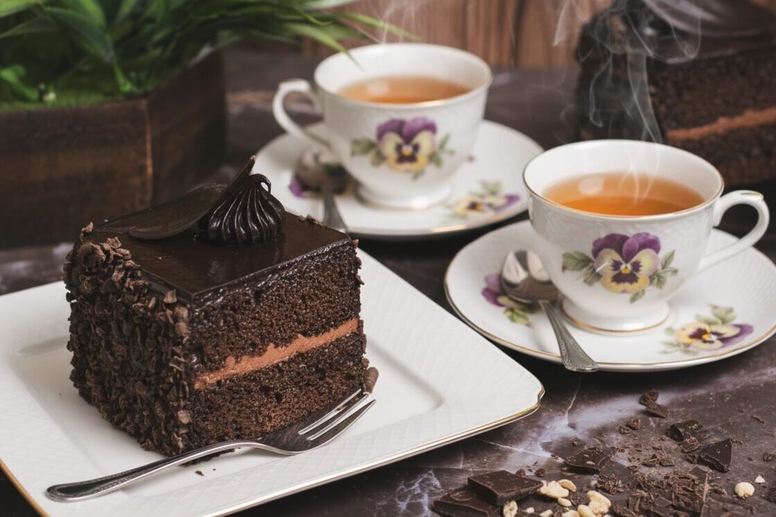 Rich chocolate cake and steaming tea in floral cups on a marble table next to chopped chocolate.