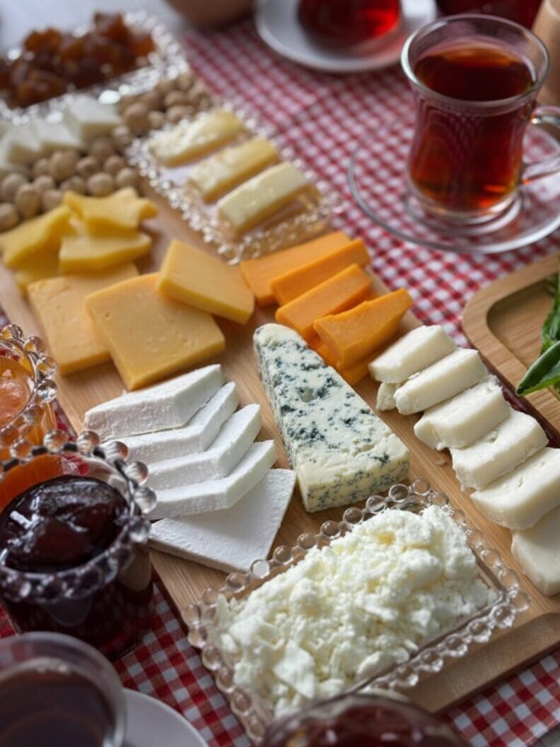 Assorted cheeses on a wooden board with jams, nuts, and tea on a red checkered tablecloth.