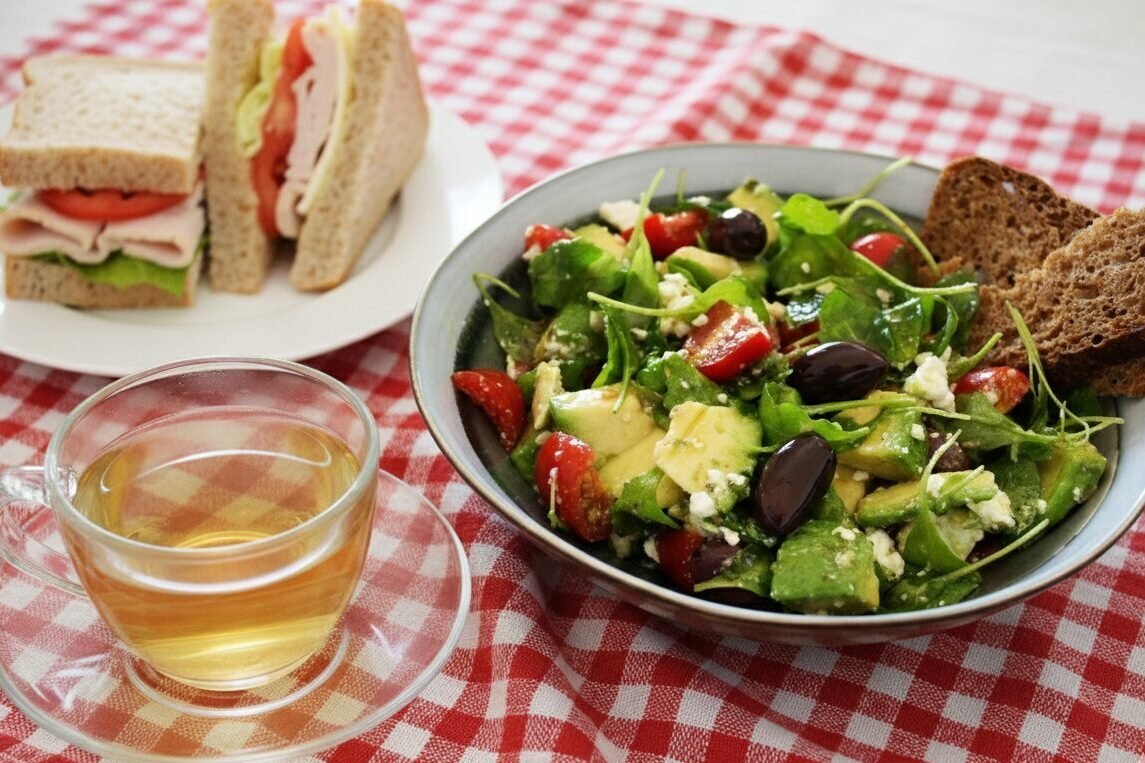 Fresh salad with tomatoes and olives, sandwich, and tea on a red checkered cloth. Perfect for a light lunch.