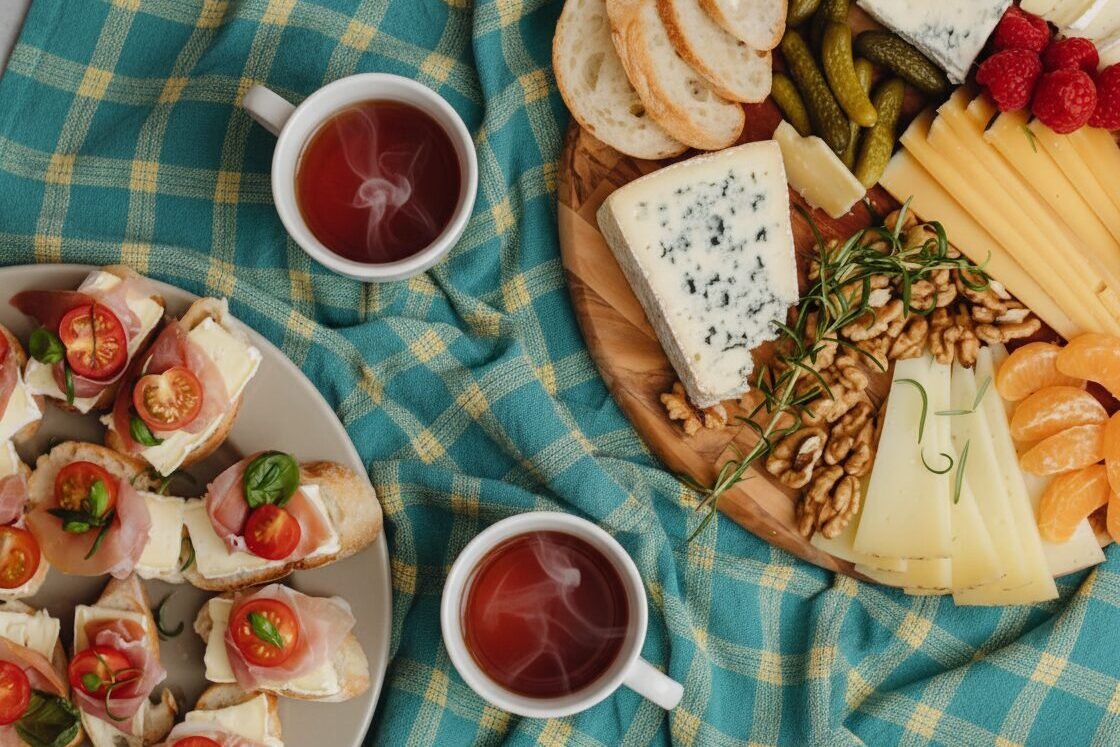 Tea with charcuterie board and open sandwiches featuring assorted cheeses, meats, nuts, and fruits on a checkered cloth.