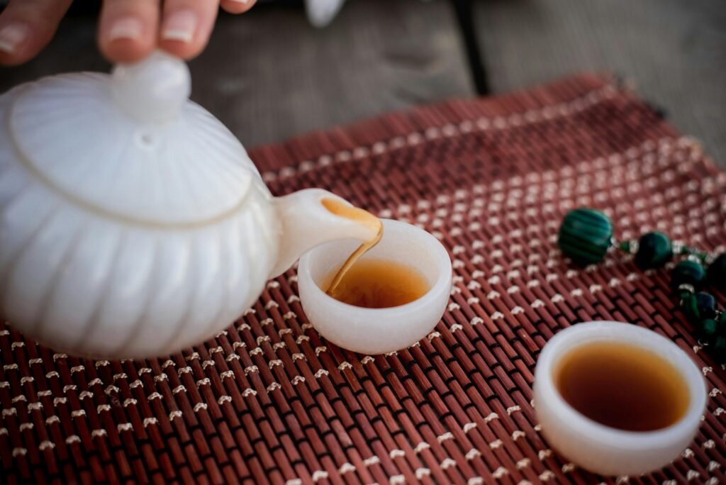Pouring tea from a white teapot into delicate cups on a woven mat with beads nearby.