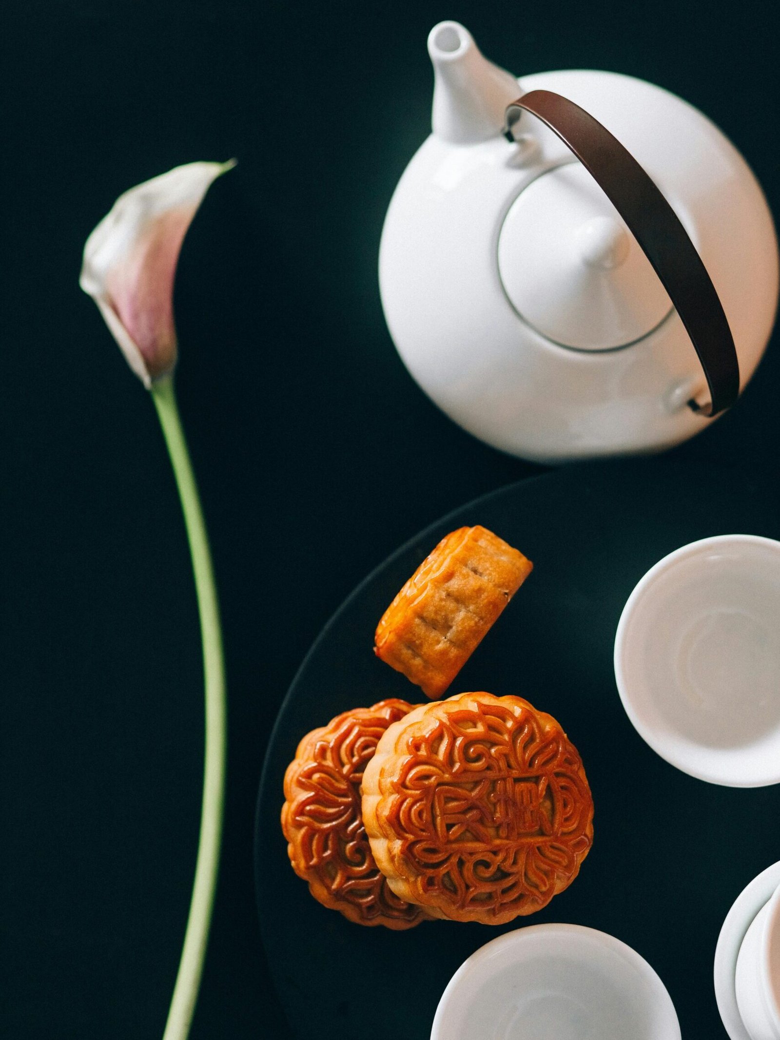White teapot with mooncakes and teacups on a dark background beside a single calla lily. Perfect for a tea setting.