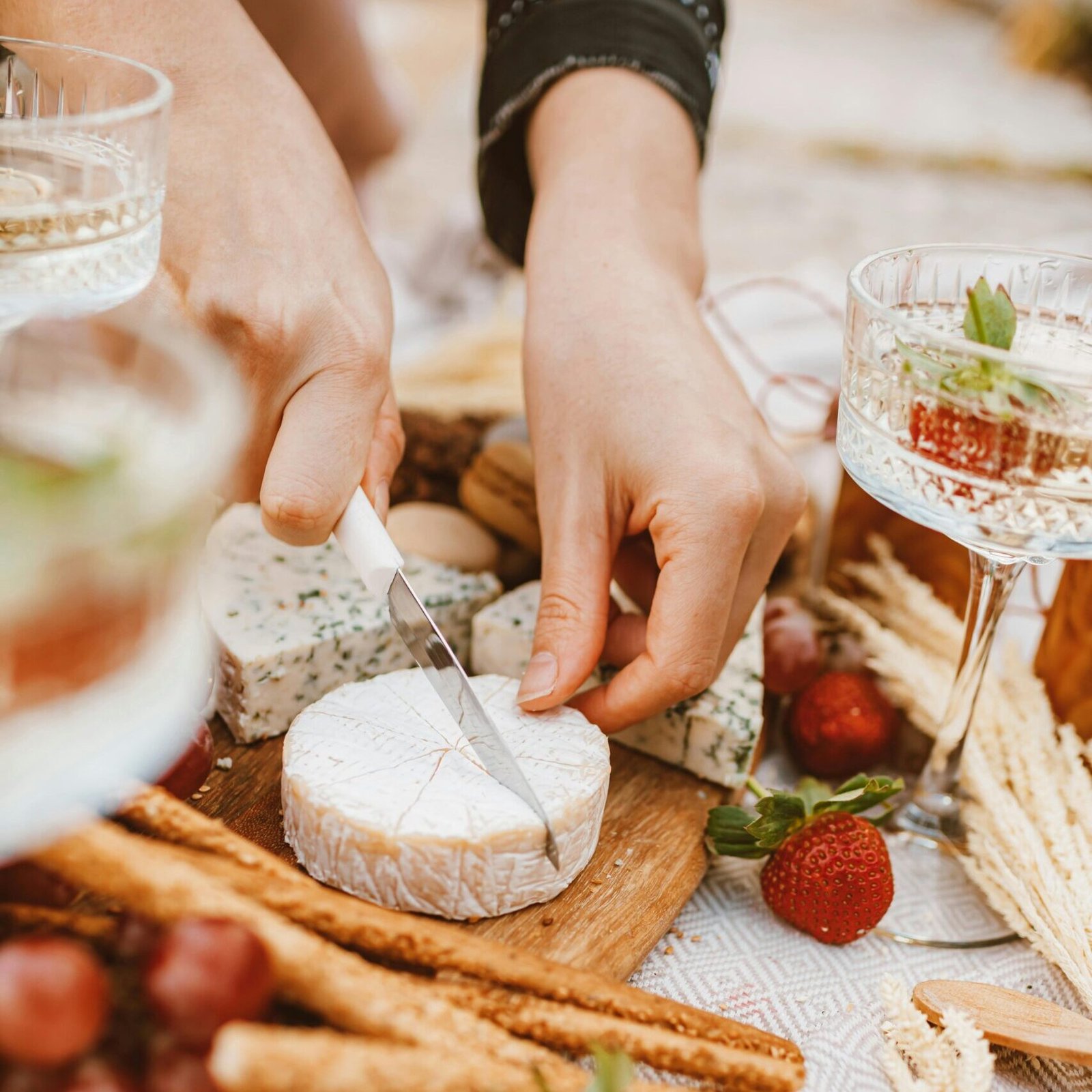 Cutting cheese on a picnic platter with fruits, breadsticks, and drinks on a decorative tablecloth.