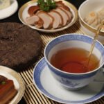 Traditional Chinese meal with tea, roasted duck, dumplings, pork belly, cheese, and chocolate on a bamboo mat.